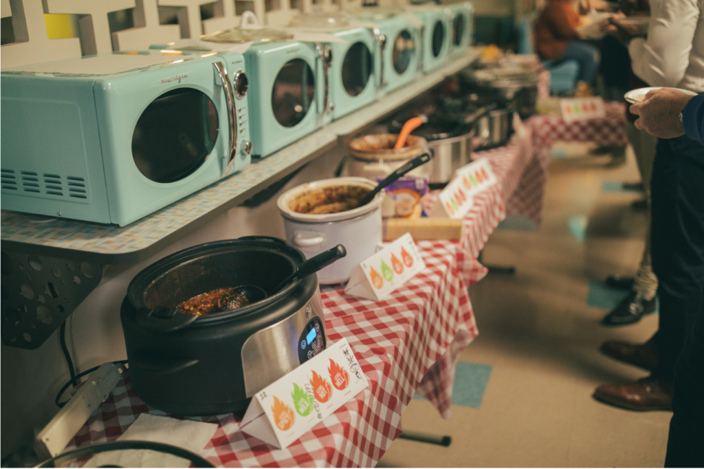 Buffet table with slow cookers and a variety of dishes, set on checkered tablecloths with spice level indicators.