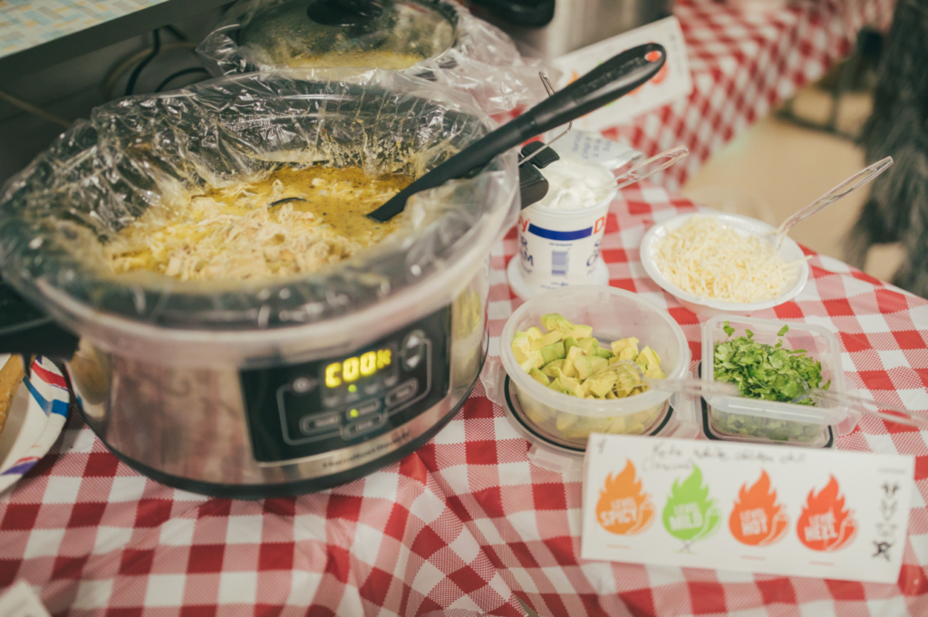 Slow cooker with white chicken chili on a checkered tablecloth, surrounded by toppings like avocado, cilantro, cheese, and sour cream.