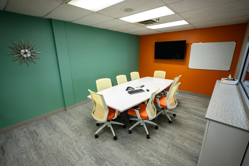 Modern conference room with a large white table, eight orange and white chairs, a teal wall on the left, an orange wall with a flat-screen TV and whiteboard on the right, and a gray patterned carpet floor.