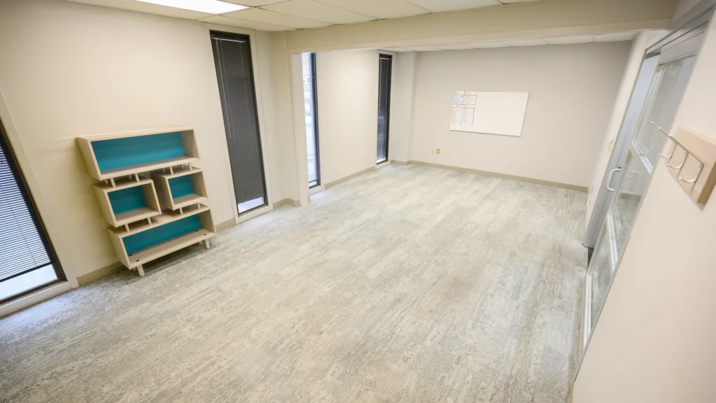 An empty office room with beige walls, carpeted floor, a teal-accent bookshelf in the corner, windows with blinds, and a glass door.