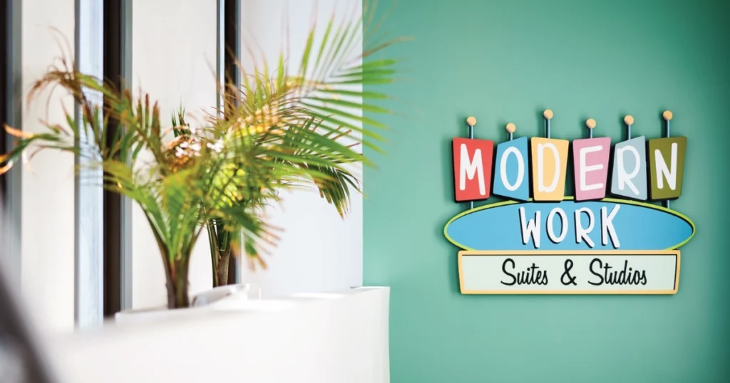 Colorful sign saying "MODERN WORK" in block letters above "Suites & Studios" on a teal background beside an out-of-focus plant on a white surface.