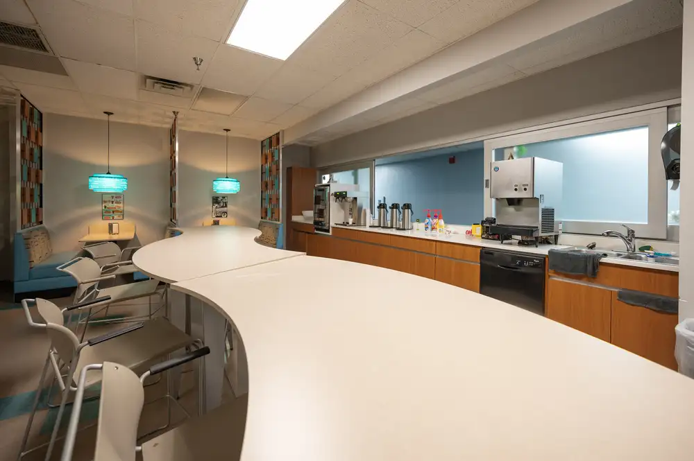 Modern office kitchen with curved countertop, beige high chairs, blue booth seats, turquoise pendant lights, and various kitchen appliances.