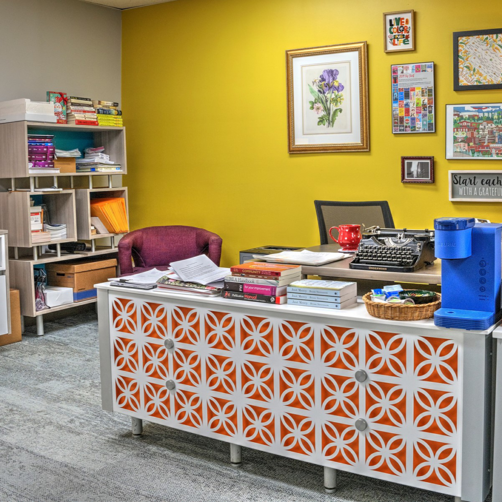 An office space with a bright yellow wall featuring framed art and a typewriter on a white patterned desk with books, a red mug, and a water dispenser nearby, conveying a creative and personalized work environment.