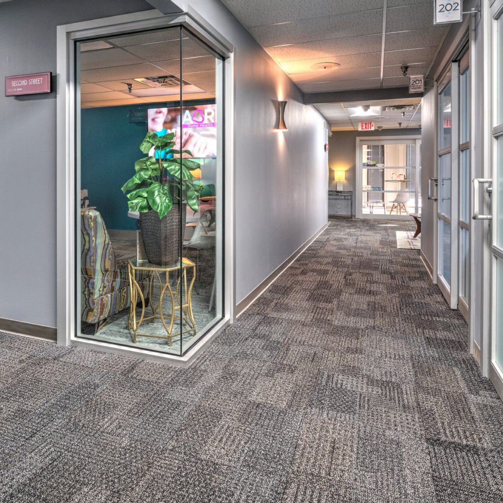 An indoor corridor in a commercial building with light blue doors, frosted glass windows, carpeted floors, and a lit exit sign at the far end.