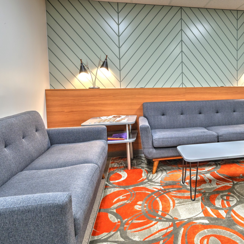 Modern lounge area with a dark gray tufted sofa, a geometric teal patterned wall, a vibrant orange and red patterned carpet, and contemporary furnishings.