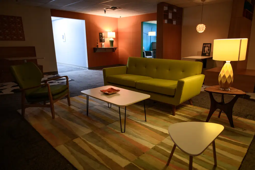 Co-working office space with a green sofa, armchair, and coffee table on a colorful rug, surrounded by orange and white walls, lamps, and various decorations.