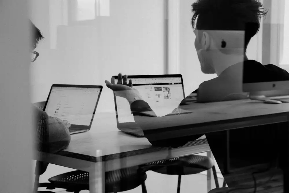 Two people working on laptops at a wooden table in a glass-walled co-working space.