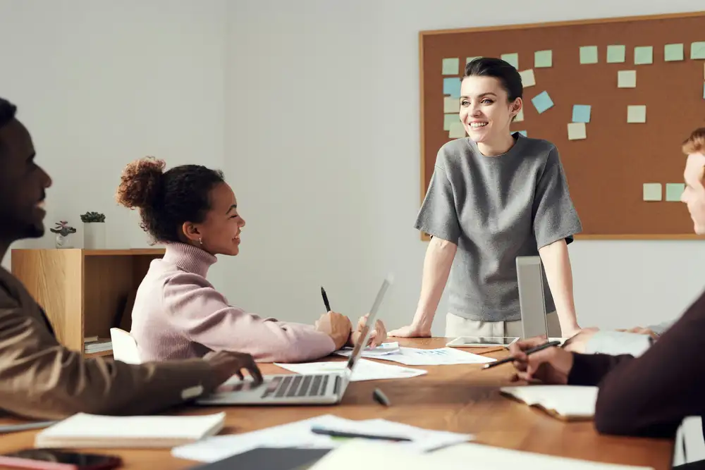 Four people in a meeting at an office table with laptops and notebooks; one person is standing and smiling.