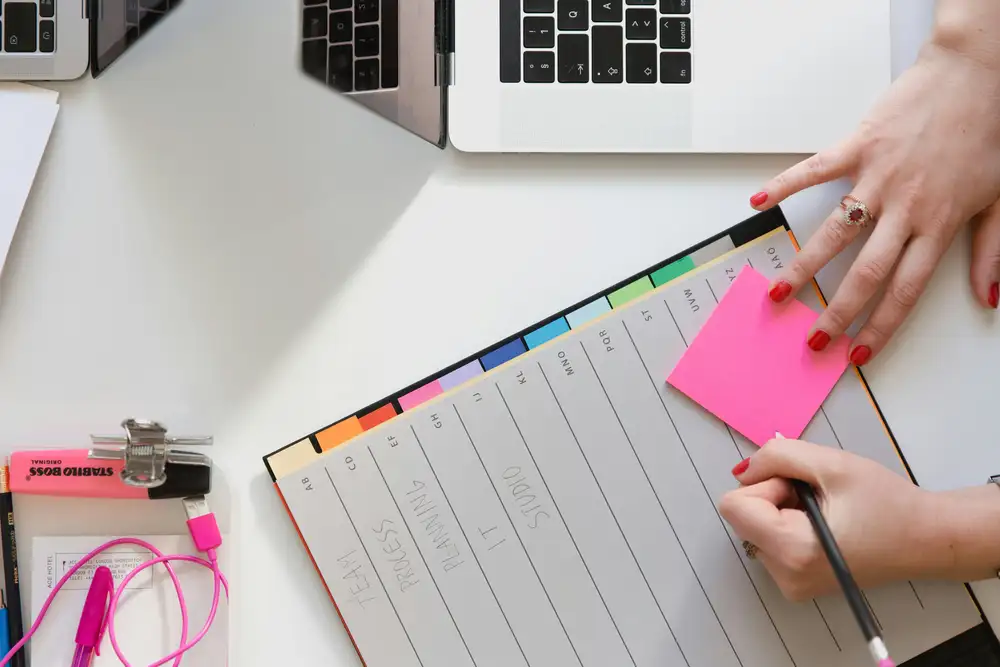 Workspace with laptops, planner, and office supplies, including a person placing a pink sticky note on the planner.