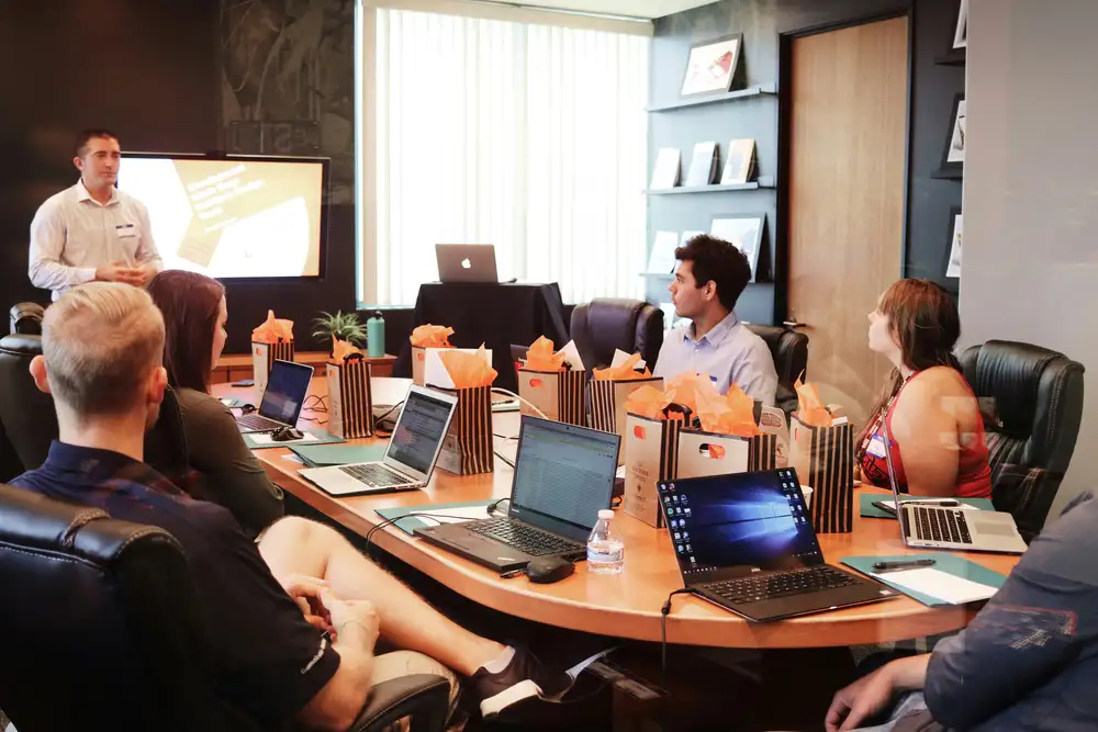 People in a meeting room with laptops and gift bags on the table, focusing on a presenter pointing to a presentation slide.