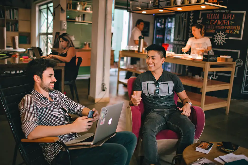 Two men sitting and conversing in a modern coworking space, with a laptop and smartphone. Additional individuals in the background working at desks. Chalkboard menu and relaxed modern decor around.