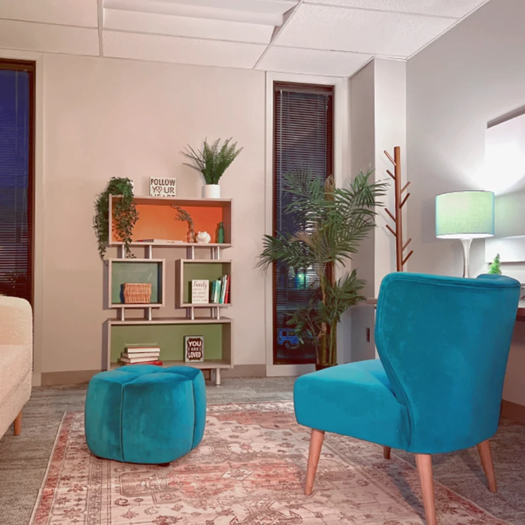 Modern living room corner with a shelving unit holding plants and decorative items, flanked by windows with blinds, a teal armchair with ottoman on a patterned rug, and a glowing table lamp.