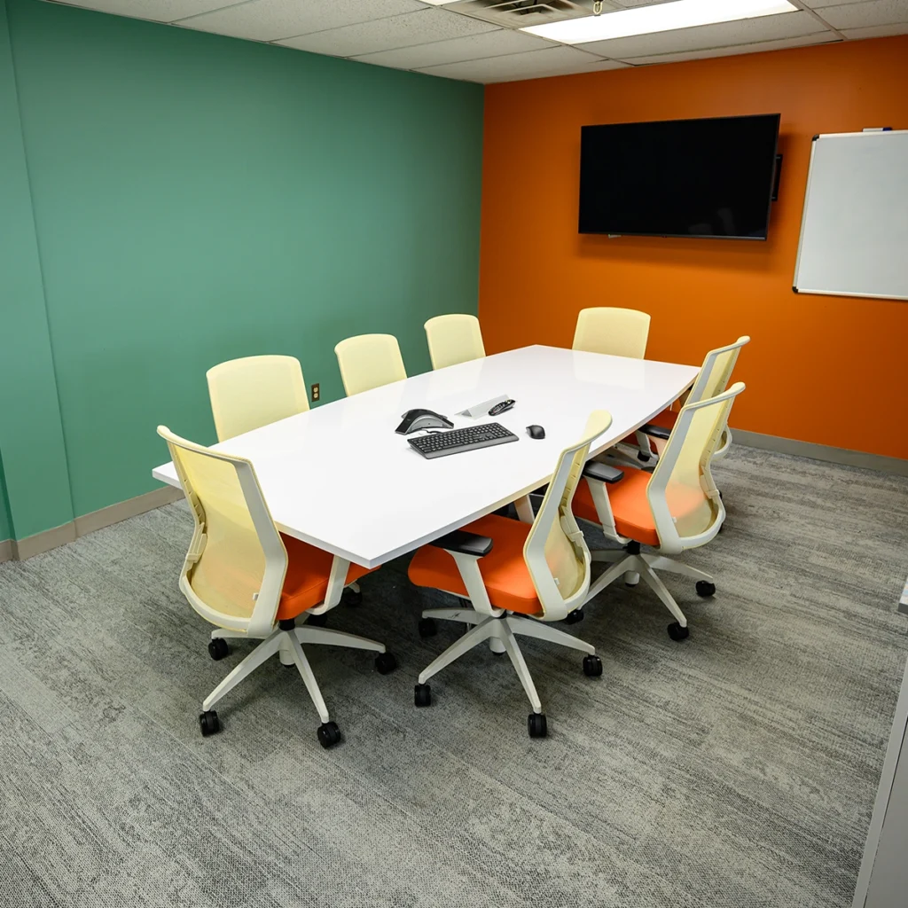 Modern conference room with a large white table, eight orange and white chairs, a teal wall on the left, an orange wall with a flat-screen TV and whiteboard on the right, and a gray patterned carpet floor.