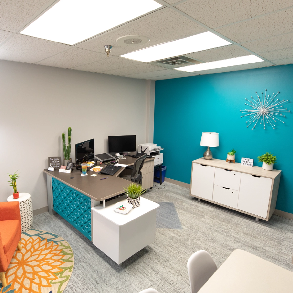 A neatly organized modern office with an L-shaped desk, teal accent wall, decorative starburst art, and white furniture accents under bright fluorescent lighting.