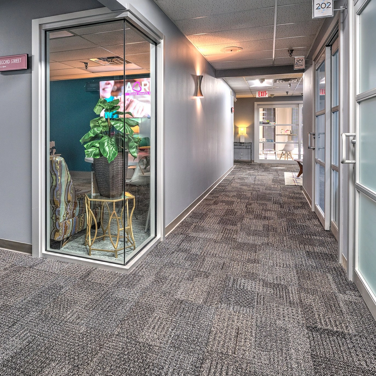 An indoor corridor in a commercial building with light blue doors, frosted glass windows, carpeted floors, and a lit exit sign at the far end.