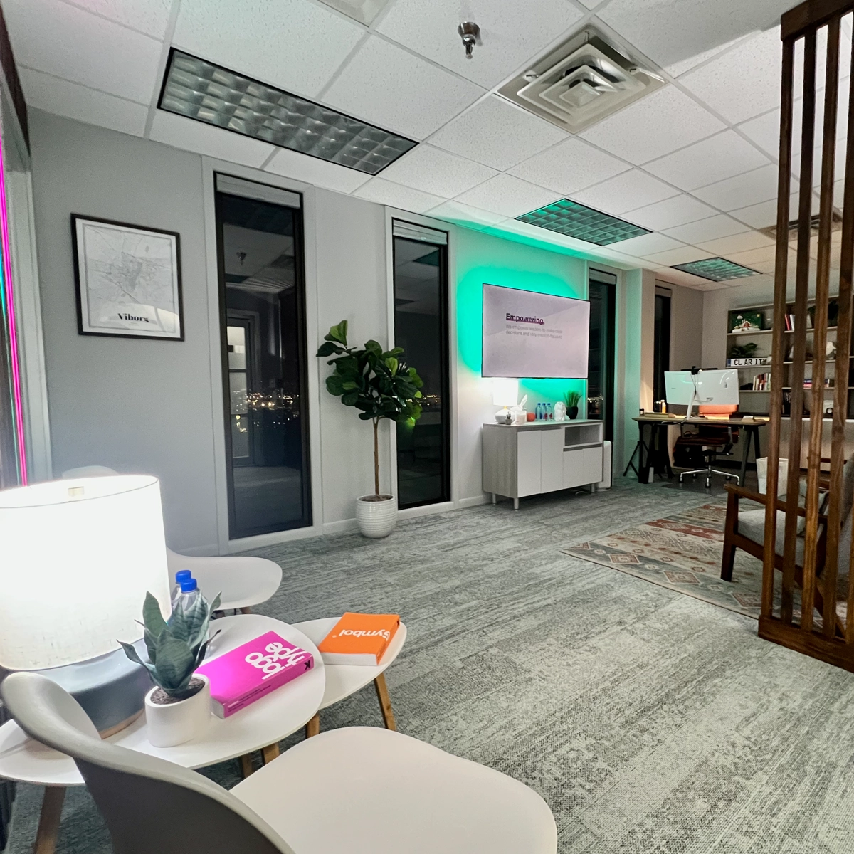 Modern office with seating area, LED-lit "Empowering" sign on monitor, workstation with dual monitors, and decorative accents like plants and colorful notebooks.