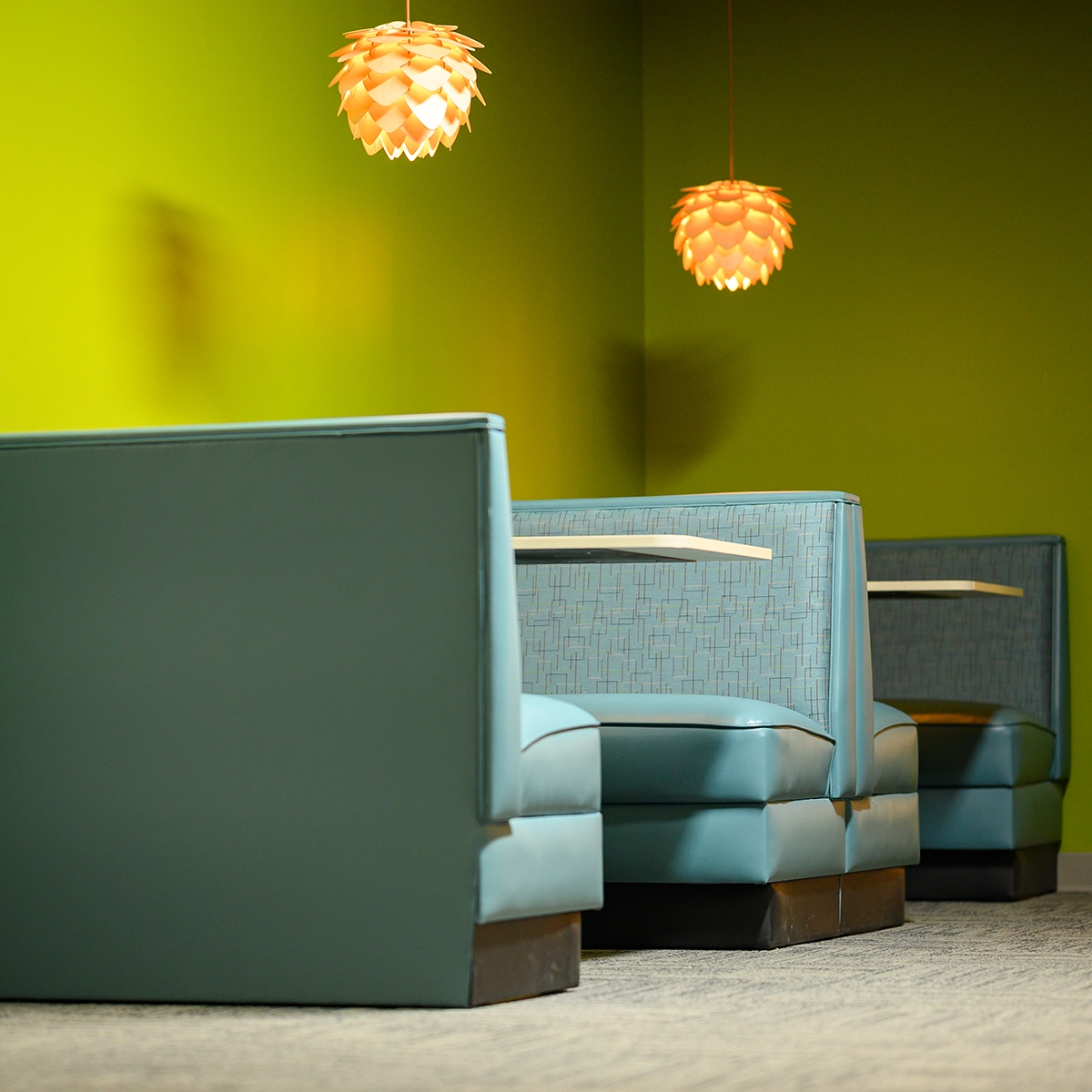 Two turquoise high-backed armchairs against a gradient green-yellow wall with warm orange pendant lights hanging above in a modern interior.