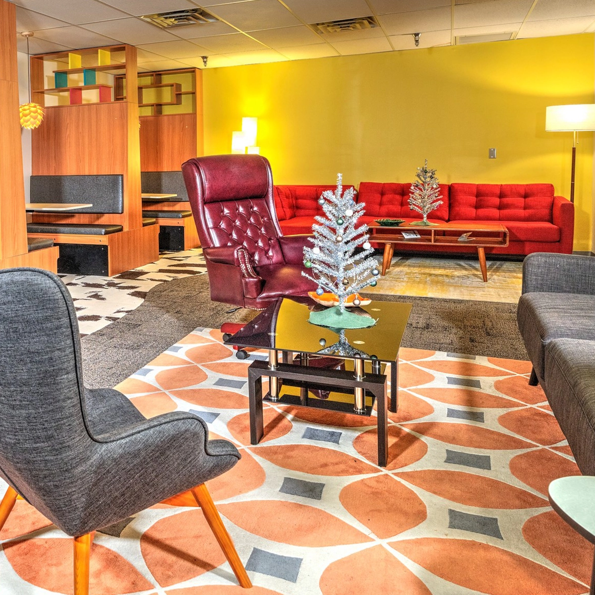 A retro-inspired living room featuring a gray chair, a red sectional sofa, a silver artificial Christmas tree on a coffee table, with mustard-yellow walls and a colorful patterned rug.