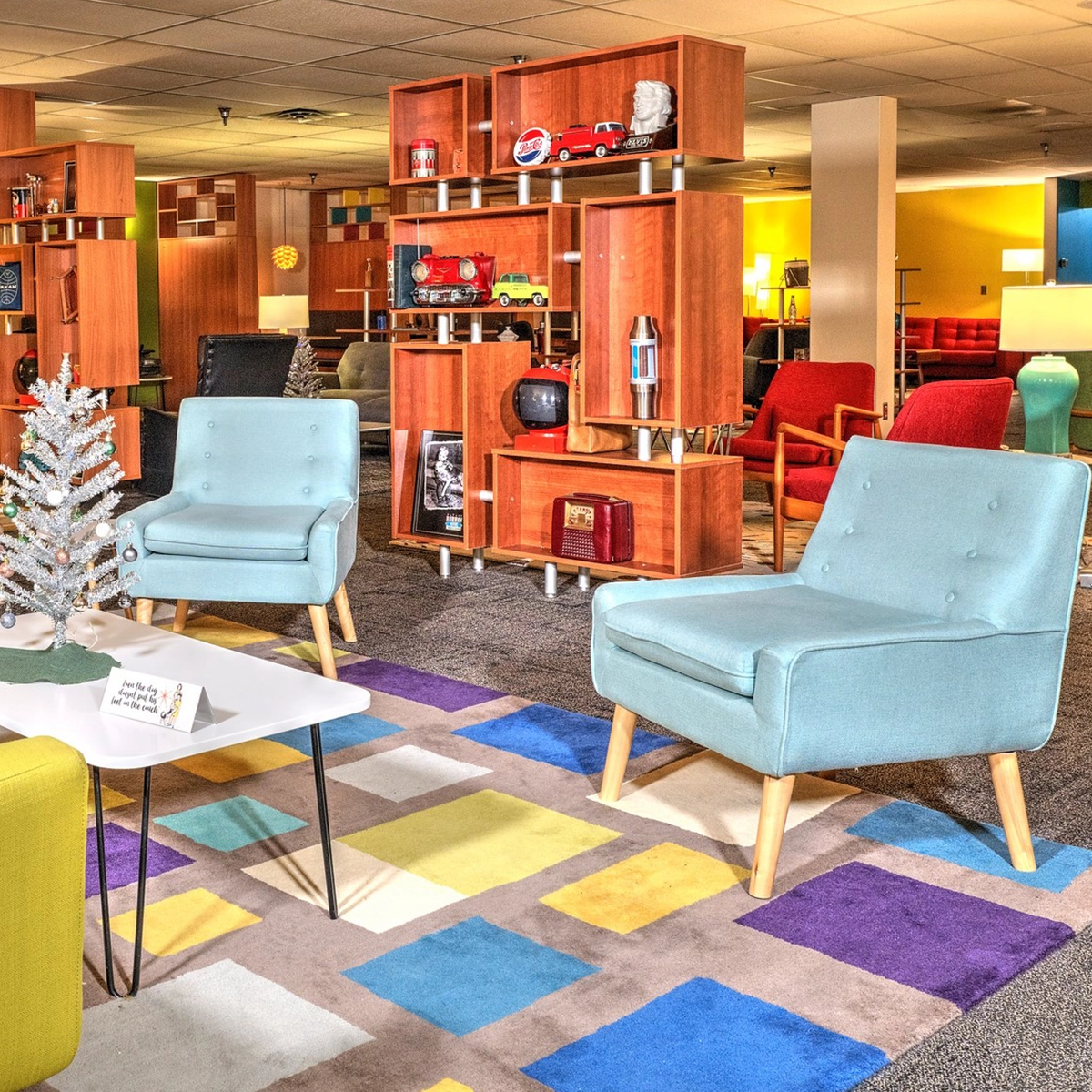 A mid-century modern styled lounge with colorful furniture, decorative shelving displaying classic items, and a multicolored geometric rug.