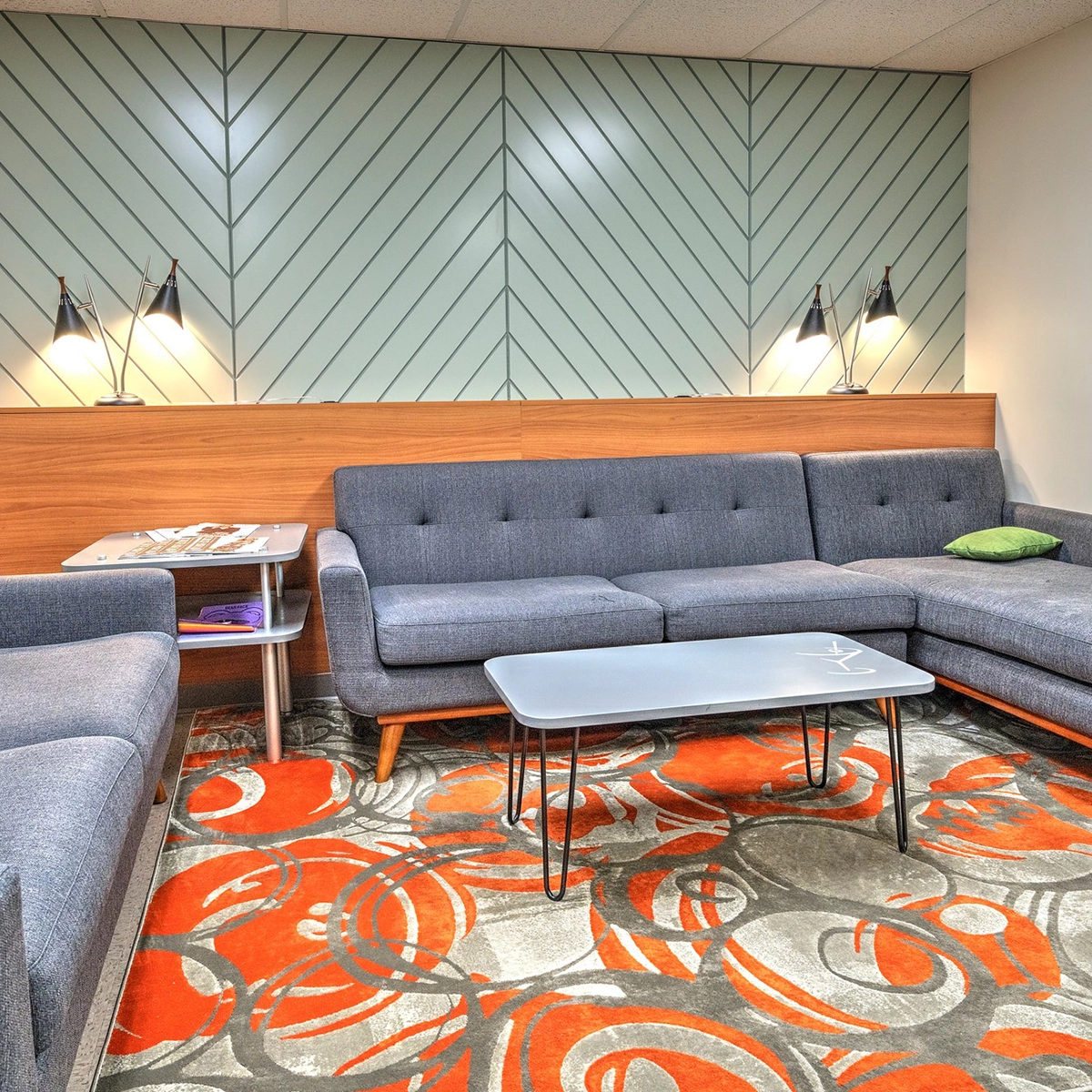 Modern lounge area with a dark gray tufted sofa, a geometric teal patterned wall, a vibrant orange and red patterned carpet, and contemporary furnishings.