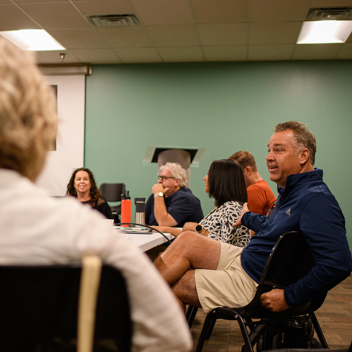 A group of six adults participating in a discussion within a meeting room with green walls. A man in a navy blue jacket appears centered in the composition, engaged with surrounding attendees.