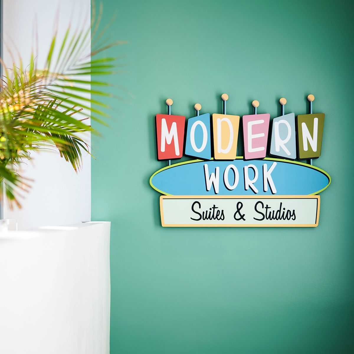 Colorful sign saying "MODERN WORK" in block letters above "Suites & Studios" on a teal background beside an out-of-focus plant on a white surface.