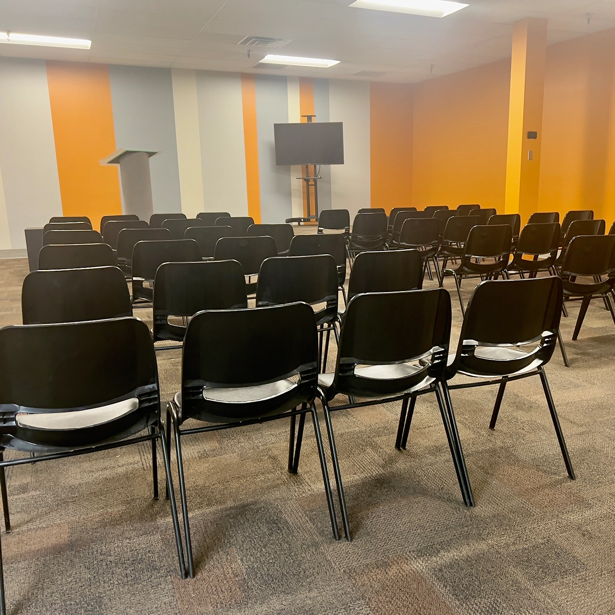 An empty auditorium with rows of black chairs facing a flat-screen TV and lectern, set against a wall with orange and gray stripes.