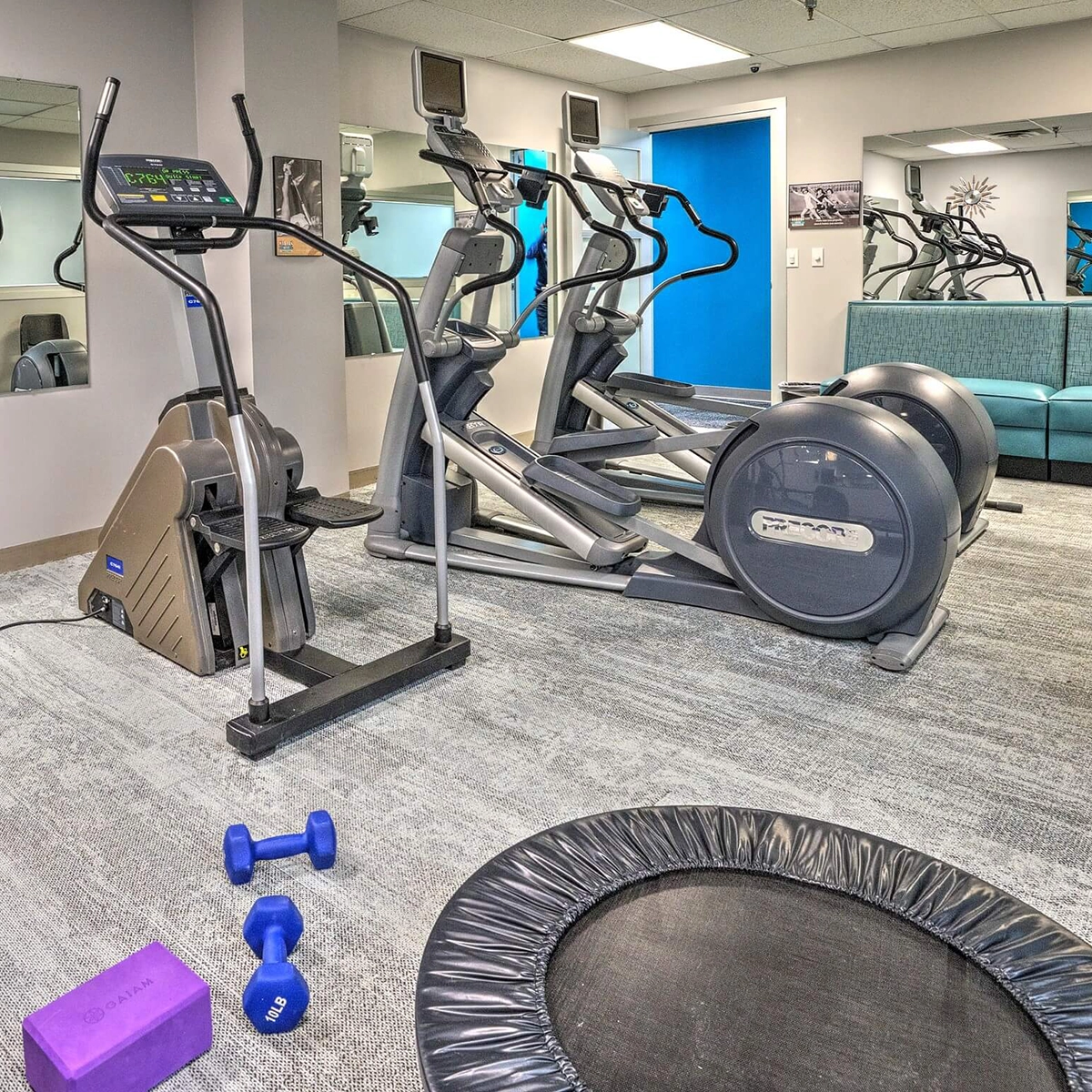 Low Impact Gym featuring elliptical machines, a stationary step machine, a small trampoline, and light hand weights with a yoga block, all set in a modern room with mirrors and a blue accent wall.
