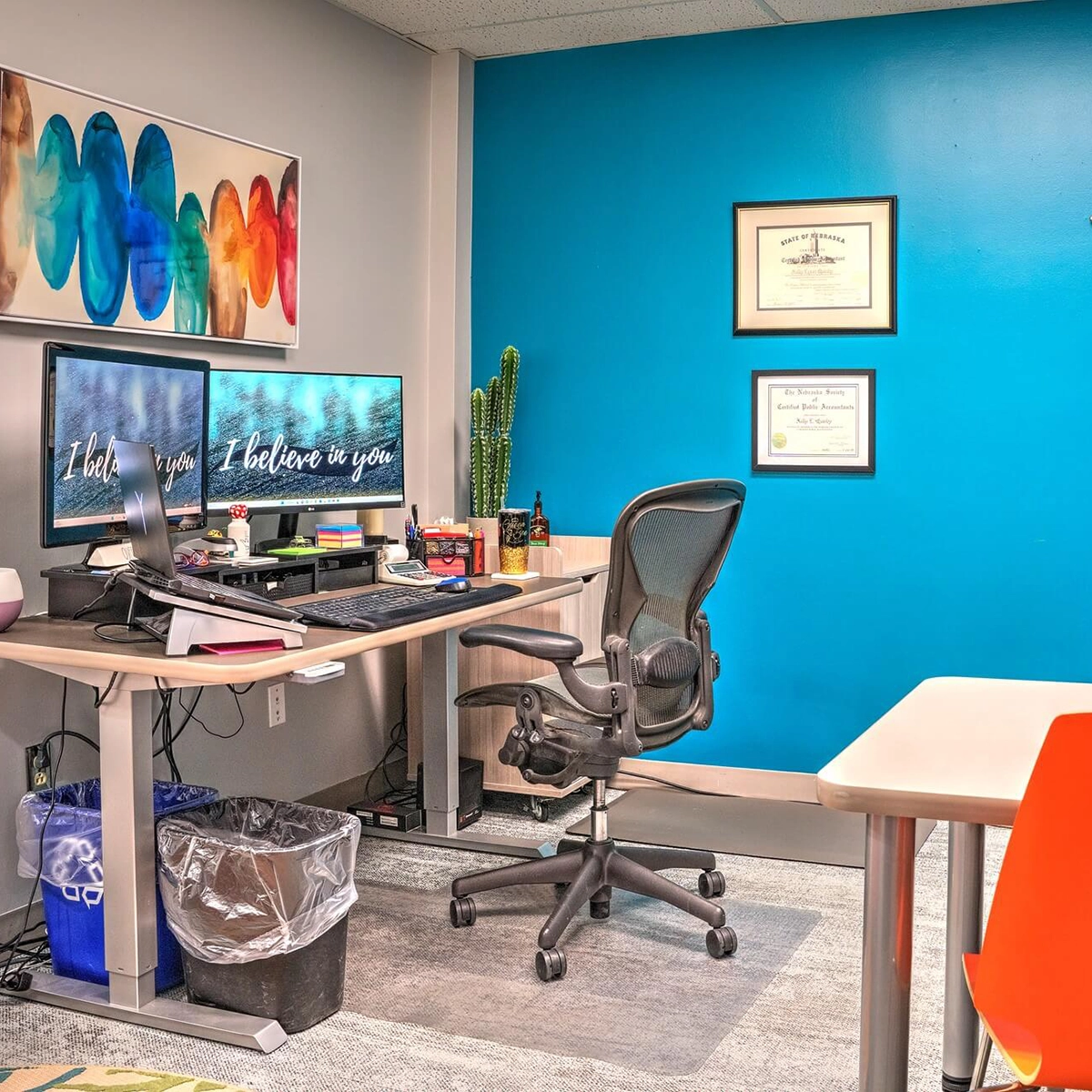 A bright office space with a turquoise blue wall, a desk with a dual monitor setup displaying "I believe in you," an ergonomic chair, a cactus plant, framed certificates, and colorful abstract art.