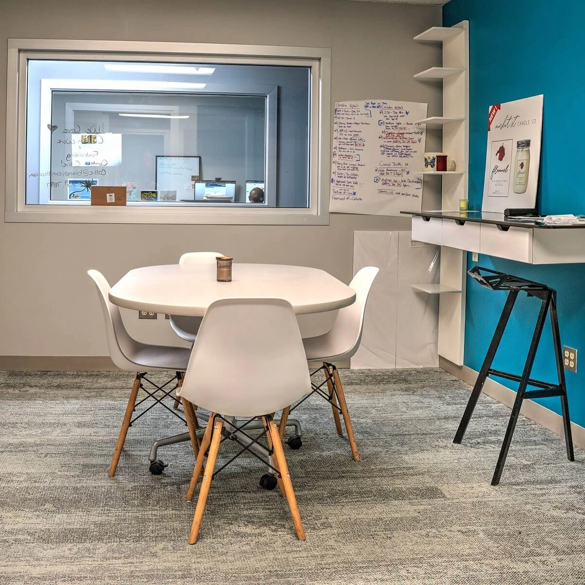 Modern office meeting area with a round white table, four chairs, a textured gray carpet, teal and white walls, a large interior window, whiteboard, and shelves with office items.