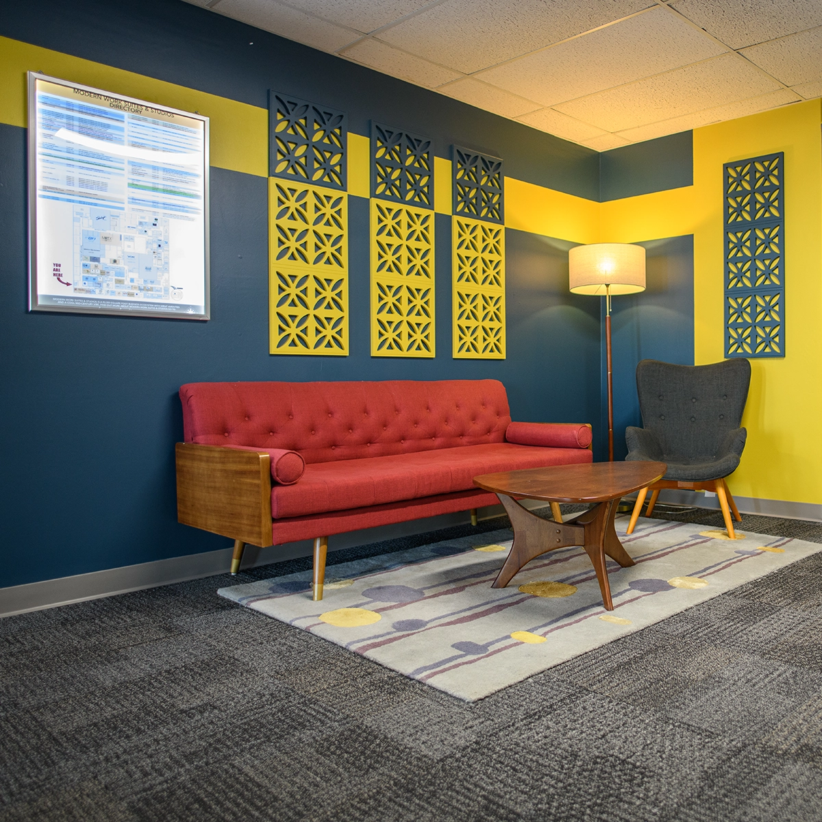 Modern mid-century style lounge with a red tufted sofa, grey armchair, wooden coffee table, and decorative geometric wall panels against blue and yellow walls.