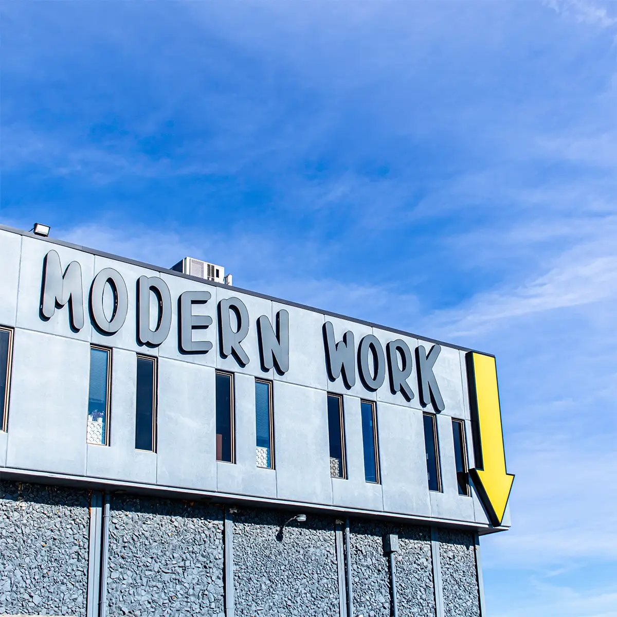 A building with Modern Work in bold letters and a large yellow arrow on the side, set against a clear blue sky, hints at dynamic office space ready to inspire productivity and innovation.